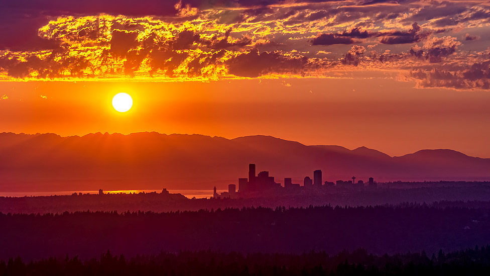 The sun sets over a city skyline with mountains in the background and scattered clouds in an orange and purple sky. Evergreen State Heat & AC