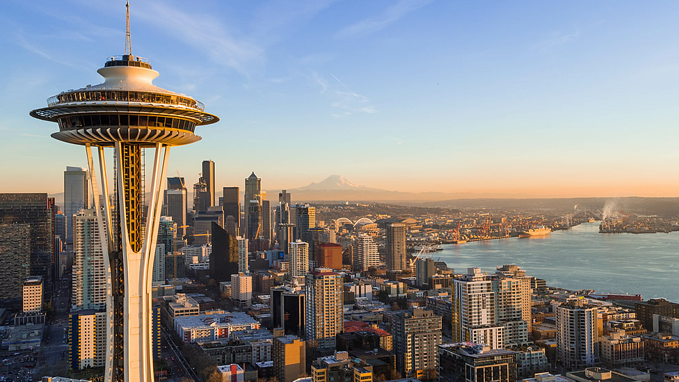 The Space Needle stands in the foreground with downtown Seattle skyscrapers behind, Mount Rainier in the distance, and Elliott Bay to the right under a clear sky. Evergreen State Heat & AC