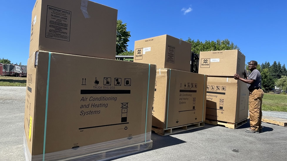 A man stands next to several large cardboard boxes labeled "Air Conditioning and Heating Systems" outdoors on a sunny day. Evergreen State Heat & AC