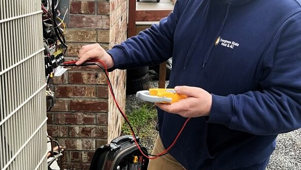 A person in a blue hoodie uses a multimeter to test the wiring on an outdoor HVAC unit next to a brick wall. Evergreen State Heat & AC