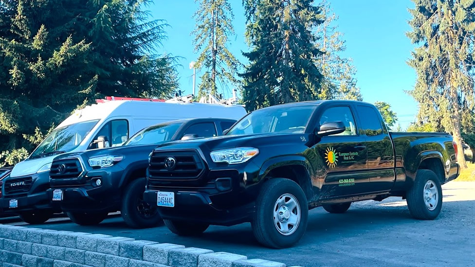 Three vehicles, including a van and two pickup trucks, are parked in a lot by a row of trees. The black truck has a yellow sun and green logo on its door. Evergreen State Heat & AC