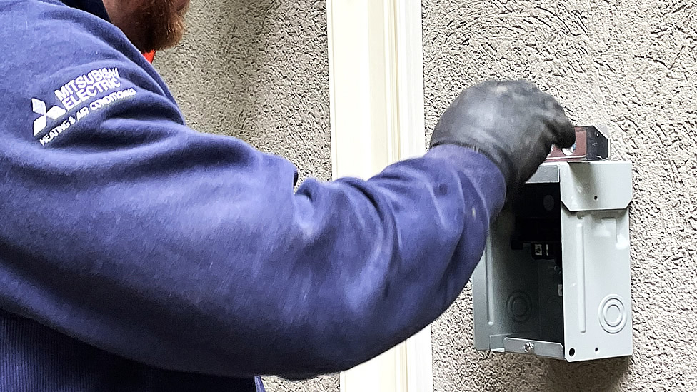 Person in a blue Mitsubishi Electric jacket and black glove works on an outdoor electrical breaker box mounted on a textured wall. Evergreen State Heat & AC
