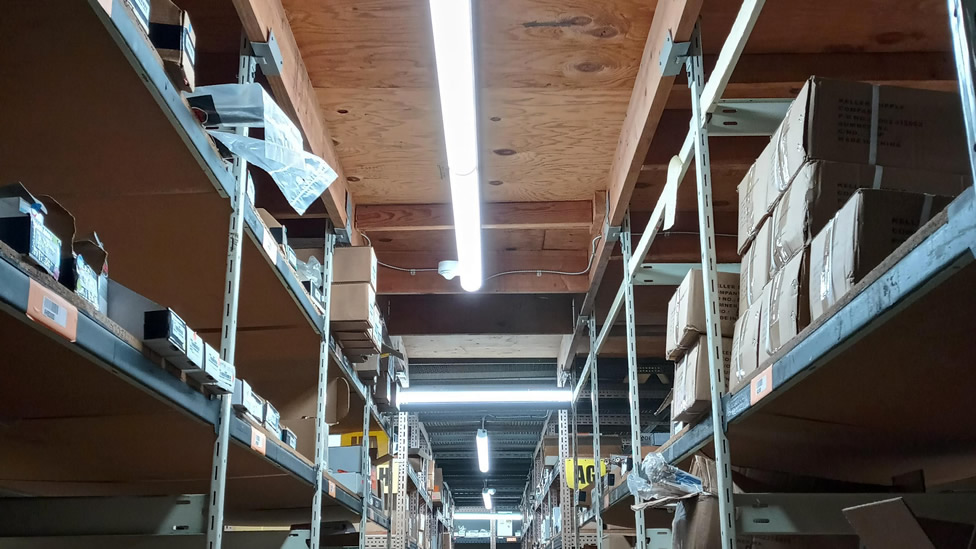 Metal shelves with assorted labeled boxes and items are arranged under wooden ceiling panels and fluorescent lighting in a storage warehouse. Evergreen State Heat & AC