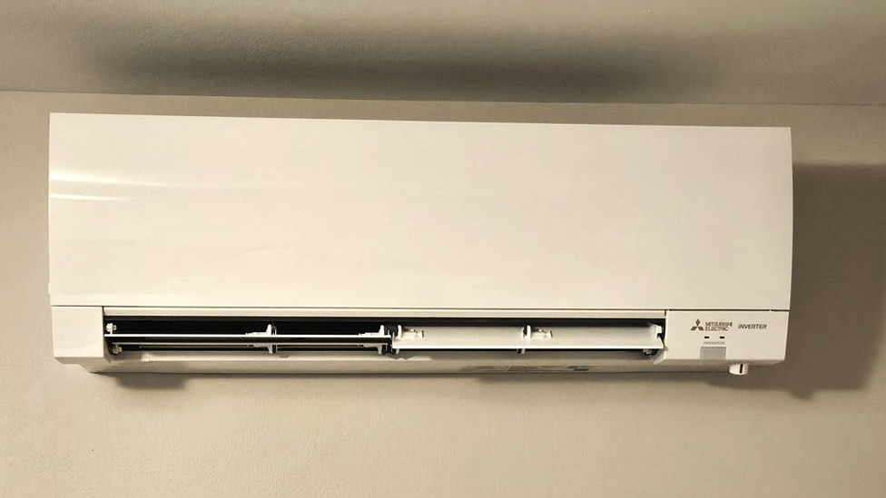 Wall-mounted Mitsubishi electric air conditioner unit with front vent partially open, installed on a plain beige wall, showcasing the sleek design commonly found in ductless heat pumps. Evergreen State Heat & AC