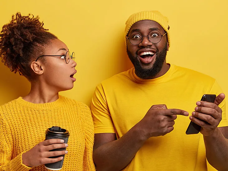 Two people in yellow sweaters stand against a yellow background. One holds a coffee cup, while the other points at a smartphone—both appear surprised, as if they've just discovered how easy it is to make a referral. Evergreen State Heat & AC