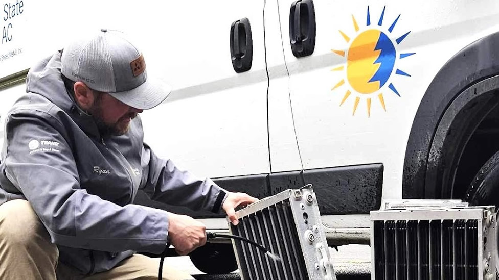 A technician in a gray jacket cleans an AC unit coil outdoors near a white van with a sun and snowflake logo, ensuring optimal indoor air quality. Evergreen State Heat & AC