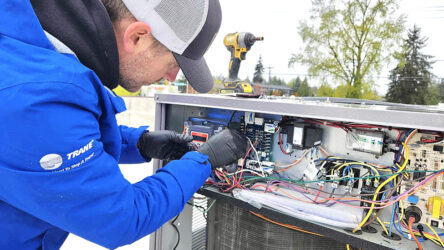 A technician in a blue jacket and black gloves repairs the wiring inside an HVAC unit outdoors, with tools and colorful wires visible. Evergreen State Heat & AC