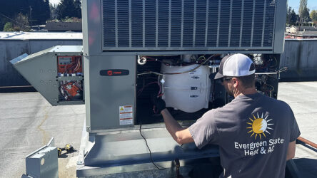 A technician wearing an "Evergreen State Heat & AC" shirt services a rooftop HVAC unit under daylight. Evergreen State Heat & AC
