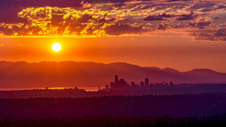 The sun sets over a city skyline with mountains in the background and scattered clouds in an orange and purple sky. Evergreen State Heat & AC