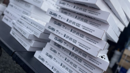Stacked white boxes labeled "Columbia GPF HP8 16202" sit on a table outdoors, with some boxes slightly askew and background objects blurred. Evergreen State Heat & AC