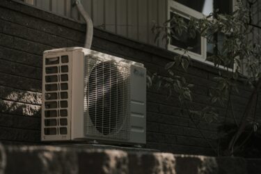 Outdoor air conditioning unit installed against a brick wall, partially shaded by a nearby tree, with a pipe attached to the top of the unit. Evergreen State Heat & AC