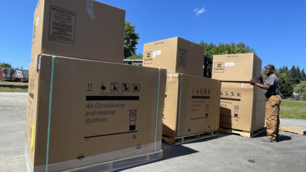 A man stands next to several large cardboard boxes labeled "Air Conditioning and Heating Systems" outdoors on a sunny day. Evergreen State Heat & AC