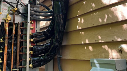 Close-up of insulated HVAC refrigerant lines connected to an outdoor unit mounted on the side of a house with horizontal wooden siding. Evergreen State Heat & AC