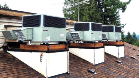 Three large HVAC units are mounted on wooden platforms atop a shingled roof, with trees and a partly cloudy sky in the background. Evergreen State Heat & AC