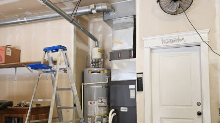 A garage corner with a step ladder, water heater, HVAC unit, metal ductwork, shelves with boxes, wall fan, and a white door with a decorative sign above it. Evergreen State Heat & AC
