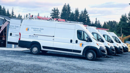 Four white service vans with "Evergreen State Heat & AC" logos are parked in a row on a gravel lot, with trees and equipment visible in the background. Evergreen State Heat & AC